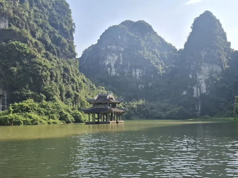 A wooden pavilion rises from calm river waters beneath towering limestone karsts in Ninh Binh.